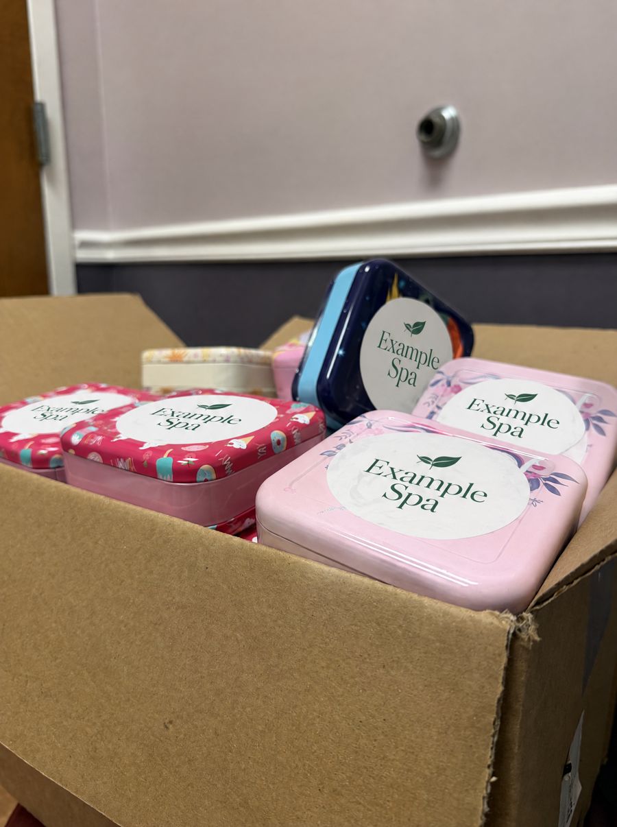 A box of colorful keepsake tins, each labeled Example Spa, packed as welcome gifts.