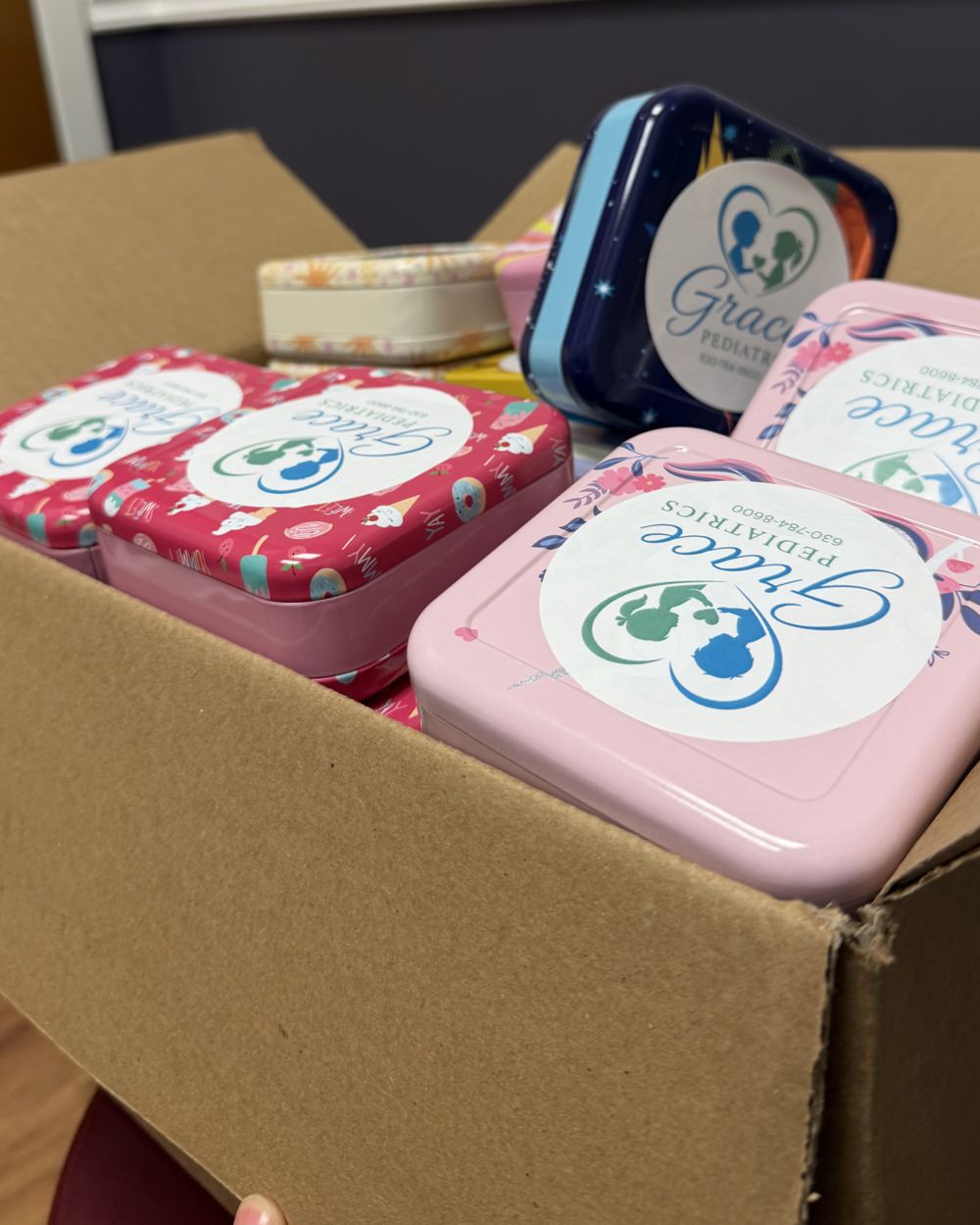 A shipping box of colorful keepsake tins, each carrying a pediatric practice's logo and phone number, packed for community outreach.